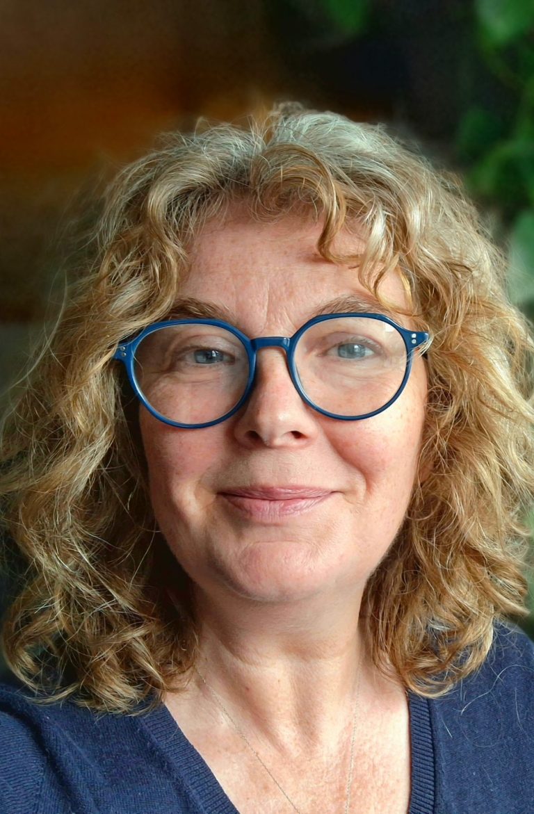 Sabine Kohlert, Portrait Frau mmit braunem Pulli und blonden mittellangen Haaren liegt mit den Armen aufgestützt auf der Wiese und schaut dabei lachend in die Kamera.