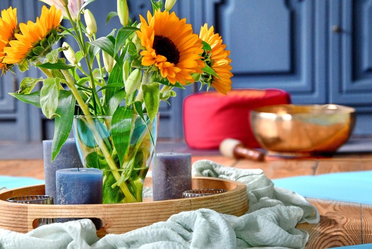 Im Meditationsraum Sonnenblumen in Glasvase auf einem Tablett mit dunkelblauen Stumpenkerzen drumherum. In Hintgergrund eine goldene Klangschale und ein rotes Meditationskissen.