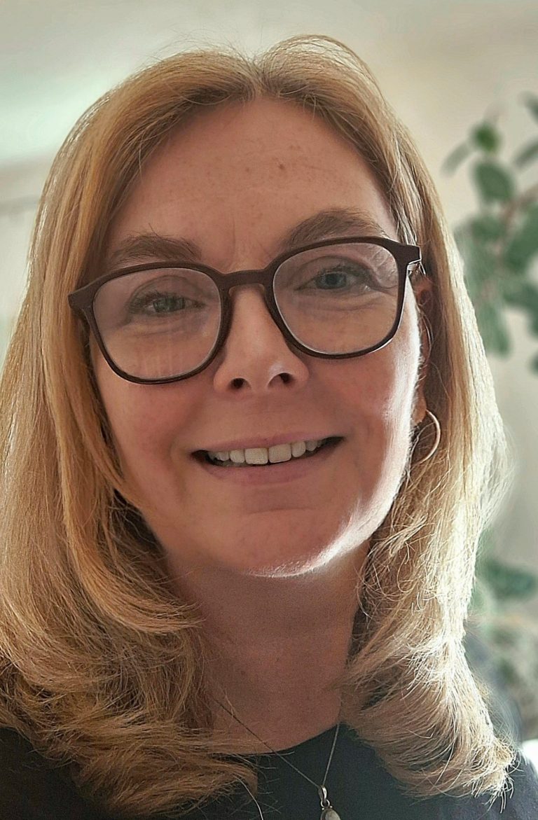 Sabine Kohlert, Portrait Frau mmit braunem Pulli und blonden mittellangen Haaren liegt mit den Armen aufgestützt auf der Wiese und schaut dabei lachend in die Kamera.