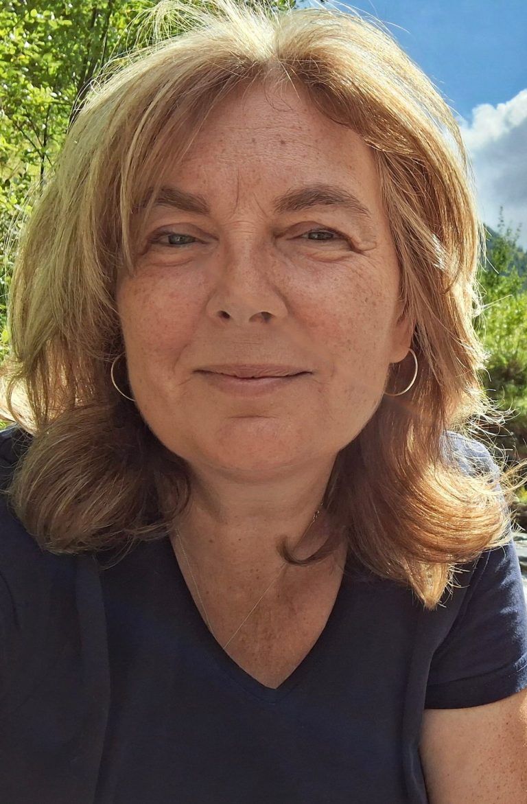 Sabine Kohlert, Portrait Frau mmit braunem Pulli und blonden mittellangen Haaren liegt mit den Armen aufgestützt auf der Wiese und schaut dabei lachend in die Kamera.