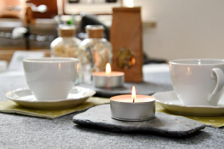 Kommen Sie gerne auf eine Tasse Tee vorbei. Zwei weiße Tassen, zwei Teelichter auf flachen Steinen. Im Hitergrund getrocknete Blumen in Glasflaschen.