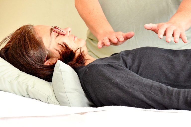 Reiki mit Conny Frau mit Brille, dunkle Haare, dunkler Pulli liegt auf Behandlungsliege. Hände einer anderen Person werden parallel über den Oberkörper gehalten.